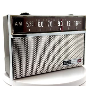 May include: A rectangular Aiwa portable radio with a dark brown casing and a silver metal speaker grille. The top section features a dark display panel with white AM frequency numbers and the text 10 TRANSISTOR. A label reads SOLID STATE AIWA.