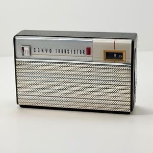 Könnte beinhalten: Ein rechteckiges Sanyo Transistorradio mit schwarzem Gehäuse, einem silbernen, perforierten Metall-Lautsprechergitter und einer silbernen Bedienblende mit dem Schriftzug SANYO TRANSISTOR, einem 6TR-Logo und einem kleinen Frequenzfenster.