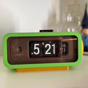 Puede incluir: Un reloj de solapa rectangular Seiko con un marco exterior de color verde lima brillante, una cara marrón y una base amarilla. La pantalla digital muestra la hora 5:21 PM con números blancos sobre azulejos negros. Un dial de alarma es visible.