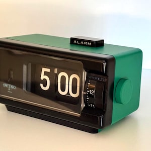Puede incluir: Un reloj de sobremesa Seiko de solapas rectangular con carcasa verde y panel frontal negro. La pantalla muestra la hora 5:00 con números blancos sobre placas negras. Un botón de alarma negro está en la parte superior y un dial en el lado derecho.
