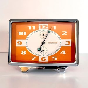 May include: A rectangular, retro-style clock with a chrome frame and base. The clock face is orange with white numerals and hands. The clock face has the text "LINDA LINDA" and a smaller dial. A classic timepiece for home decor.