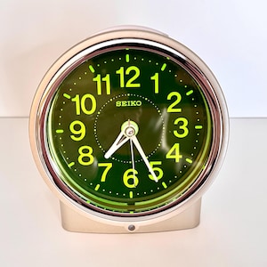 May include: A silver Seiko alarm clock with a green face and bright yellow numerals. The clock has a rounded shape and a small base. The hands are white, and the word "SEIKO" is printed on the face.