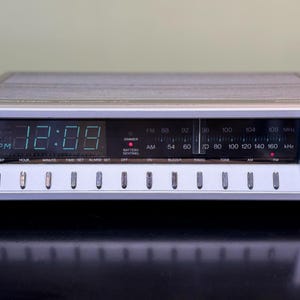 May include: Silver digital alarm clock with a green LED display showing 12:09 PM. Features include radio tuning, volume control, and various function buttons. The clock has a retro design.