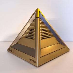 Puede incluir: Objeto dorado en forma de pirámide con acabado metálico. La pirámide tiene un diseño geométrico con ranuras de ventilación y la palabra "SEIKO" impresa en la base. La esquina superior es amarilla.