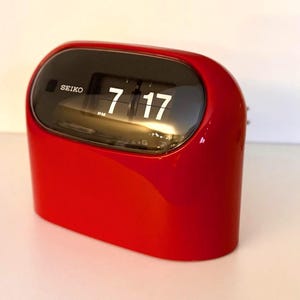 May include: A bright red Seiko flip clock with a rounded, glossy exterior. The front features a black display panel showing the time 7:17 PM in white numerals. The brand name Seiko is printed in white on the left side of the dark display window.