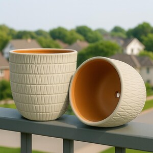 May include: Two beige ceramic plant pots with textured geometric patterns. One pot is upright, the other is tilted on its side. The interior of both pots is a warm orange colour. The pots are on a grey railing with a blurred green background.