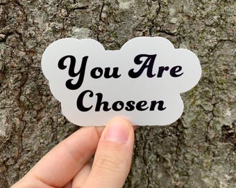 May include: A white sticker in a cloud shape with the words "You Are Chosen" in black script. The sticker is held in front of a textured tree trunk.