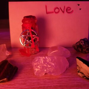 Peut inclure: Une petite bouteille en verre avec un sceau de cire rouge et un charme pentagramme argenté. La bouteille est entourée de divers cristaux et d'une carte sur laquelle est écrit le mot "Amour".