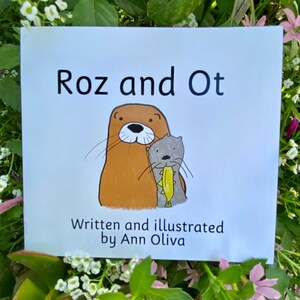 May include: A white book cover with the title "Roz and Ot" in black text. The cover features a cartoon illustration of two otters, one brown and one grey, with the grey otter holding a fish. The text "Written and illustrated by Ann Oliva" is below the illustration.