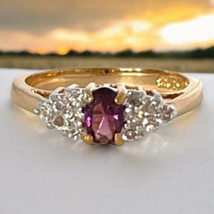 May include: A gold ring featuring a central oval-cut purple gemstone flanked by two clusters of small, clear, round-cut stones set in white metal. The band is polished yellow gold, creating a contrast with the white metal stone settings.