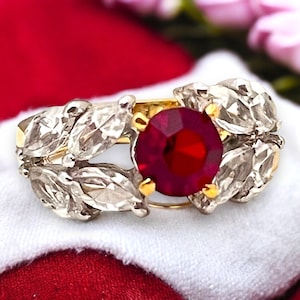 May include: A gold ring with a large red gemstone surrounded by small white gemstones set in a leaf design.