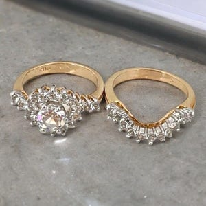 Può includere: Un set di anelli in oro composto da un anello di fidanzamento con una pietra centrale rotonda circondata da un alone di diamanti più piccoli e una fede nuziale curva abbinata con una fila di diamanti incastonati lungo il bordo superiore.