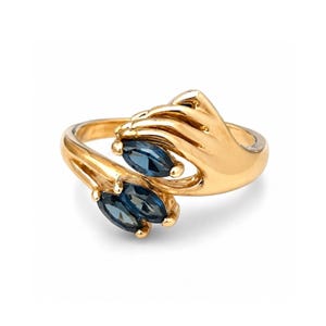 Puede incluir: Un anillo de oro con un diseño de mano esculpida que sostiene dos piedras preciosas de color azul oscuro en talla marquesa. La banda es de oro pulido y el motivo de la mano está integrado en la estructura del anillo, sujetando una piedra.