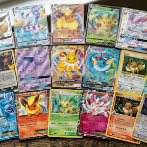 May include: A collection of Pokémon trading cards featuring various Eevee evolutions. The cards showcase vibrant illustrations of characters like Vaporeon, Jolteon, and Flareon, with holographic and foil accents. The cards are arranged on a wooden surface.