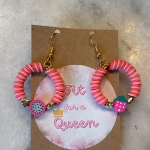 May include: Pink and red hoop earrings with gold-coloured accents. The earrings have a layered design and are adorned with fruit-shaped charms, including a dragon fruit and a strawberry. The earrings are displayed on a brown card with the text "Fit for a Queen".
