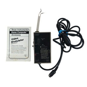 May include: A Texas Instruments Video Modulator model UM1381 with a black rectangular housing, a silver metal antenna, and a long black cable. Included is a white paper operating guide with black text detailing the home computer accessory.