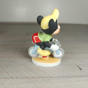 K&ouml;nnte beinhalten: Eine Mickey Mouse-Figur auf einem Fahrrad. Mickey tr&auml;gt eine gelbe M&uuml;tze, ein gr&uuml;nes Hemd, rote Shorts und gelbe Schuhe. Das Fahrrad ist hellblau. Die Figur steht auf einem cremefarbenen, runden Sockel.