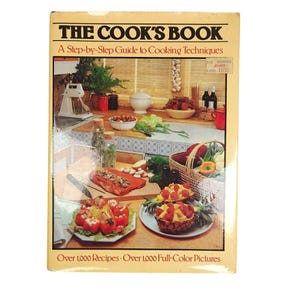Può includere: Un libro di cucina vintage intitolato "The Cook's Book" con una guida passo passo alle tecniche di cucina. La copertina presenta una scena di cucina con vari piatti, ingredienti e un robot da cucina. Il libro promette oltre 1.000 ricette e immagini a colori.