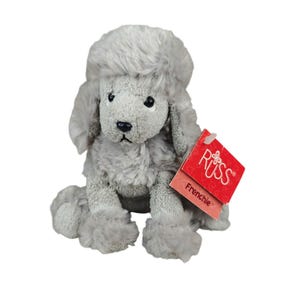 May include: A plush toy poodle with light grey, textured fur, dark eyes, and a black nose. The stuffed animal is in a seated position and features a red rectangular tag attached to its side that reads RUSS and Frenchie in white text.
