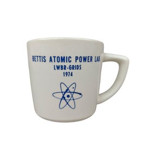 May include: A white ceramic mug featuring blue text that reads BETTIS ATOMIC POWER LAB, LWBR-GRIDS, and 1974, accompanied by a blue atomic symbol graphic. The mug has a sturdy handle on the right side and a smooth, rounded body design.