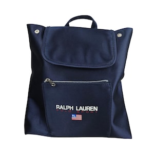 Puede incluir: Mochila azul marino con cierre de solapa y asa superior. El bolsillo delantero tiene una cremallera plateada y el logotipo "Ralph Lauren Polo Sport" con una bandera estadounidense. Diseño sencillo y clásico.