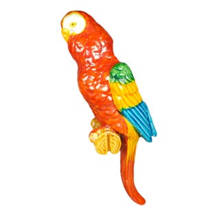 Puede incluir: Figura de loro colorido con cuerpo rojo, cabeza amarilla y alas verdes, amarillas y azules. El loro está posado en una rama amarilla. La figura es probablemente de cerámica o material similar.