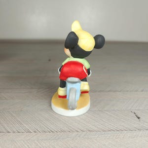 K&ouml;nnte beinhalten: Eine Keramikfigur einer Zeichentrickfigur, die ein hellblaues Motorrad f&auml;hrt. Die Figur tr&auml;gt einen gelben Helm, ein gr&uuml;nes Hemd und einen roten Sitz. Die Figur steht auf einem gelben Sockel mit wei&szlig;em Rand.