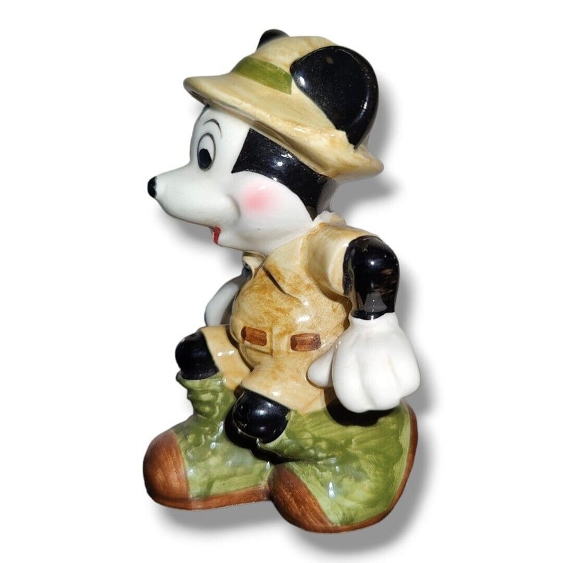 K&ouml;nnte beinhalten: Eine Keramikfigur einer Zeichentrickmaus in Safari-Outfit. Die Maus tr&auml;gt einen beigefarbenen Safarihut, eine Jacke und eine gr&uuml;ne Hose. Die Maus hat schwarze Ohren, ein wei&szlig;es Gesicht und rosige Wangen. Die Figur ist in einer hockenden Position dargestellt.