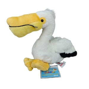 Puede incluir: Un peluche de pelícano con pelaje blanco, un gran pico amarillo y patas amarillas. Las alas tienen puntas negras. Una etiqueta azul de Webkinz está unida a la base, con el texto Webkinz discover a virtual world GANZ.