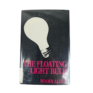 May include: A hardcover book titled "The Floating Light Bulb" by Woody Allen. The cover is black with a large white light bulb graphic and pink text. The book has a worn appearance.