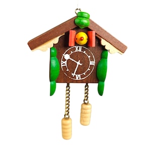 May include: A miniature wooden cuckoo clock with a brown frame, green accents, and a white clock face. The clock features a small green roof, red and yellow details, and two hanging weights. The clock is designed to be a decorative item.