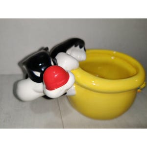Puede incluir: Un tazón de cerámica con forma de Silvestre el gato de Looney Tunes. El tazón es amarillo y presenta la cara y el cuerpo de Silvestre. Silvestre está representado con su característico pelaje blanco y negro y una nariz roja.
