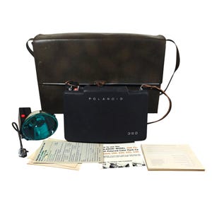 Puede incluir: Un juego de cámara Polaroid 350 Land vintage, que incluye una cámara negra, un flash y un estuche de transporte de cuero marrón oscuro. El juego también incluye manuales de instrucciones y documentos. La cámara tiene la palabra "POLAROID" impresa.