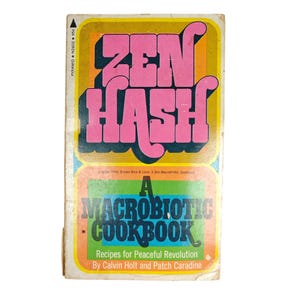 May include: Vintage paperback cookbook titled "Zen Hash: A Macrobiotic Cookbook." The cover features the title in large pink letters over a yellow and orange background. The lower section has a green and orange banner with the title "A Macrobiotic Cookbook."