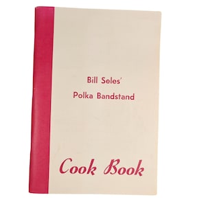 May include: A vintage cookbook titled "Bill Seles' Polka Bandstand Cook Book." The cover is cream-colored with a bright pink spine. The title is in a cursive, red font. The cookbook appears to be in good condition.