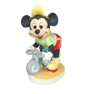 K&ouml;nnte beinhalten: Eine Keramikfigur von Mickey Mouse auf einem hellblauen Fahrrad. Mickey tr&auml;gt einen gelben Hut, ein gr&uuml;nes Hemd, rote Shorts und gelbe Schuhe. Die Figur steht auf einem beige-wei&szlig;en Sockel.