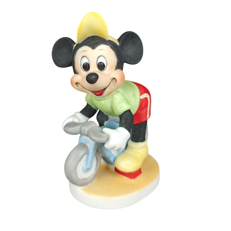 K&ouml;nnte beinhalten: Eine Keramikfigur von Mickey Mouse auf einem hellblauen Fahrrad. Mickey tr&auml;gt einen gelben Hut, ein gr&uuml;nes Hemd, rote Shorts und gelbe Schuhe. Die Figur steht auf einem beige-wei&szlig;en Sockel.