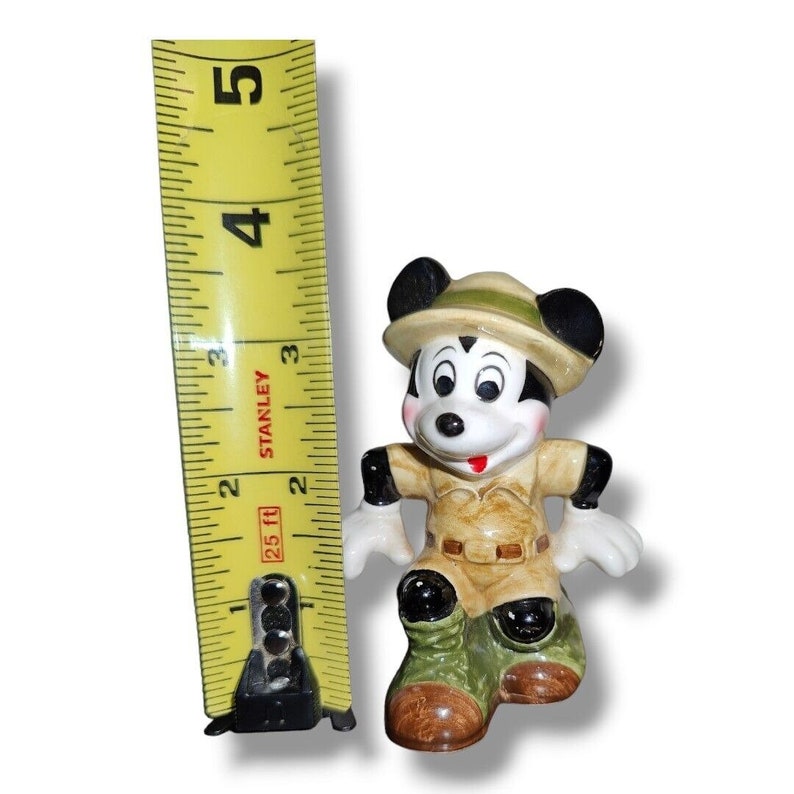 K&ouml;nnte beinhalten: Eine Keramikfigur von Mickey Mouse in Safari-Outfit, mit beigem Hut und Hemd, gr&uuml;nen Stiefeln und schwarzen Ohren. Die Figur ist etwa 7,6 cm hoch, gemessen mit einem gelben Stanley-Ma&szlig;band.