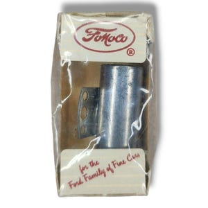 May include: A vintage car part in its original packaging. The box is tan with the "FoMoCo" logo in red. The part is a silver metal component, likely for a Ford vehicle. Text on the box reads "for the Ford Family of Fine Cars."