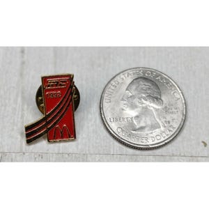 May include: A red and gold enamel pin featuring the text "ARE 1992" and the McDonald's logo. The pin has black and red stripes and is next to a quarter for size comparison.