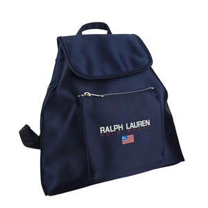 May include: Navy blue backpack with a flap closure and a small zippered front pocket. The front pocket features the text "RALPH LAUREN POLO SPORT" and a small American flag. The backpack has a top handle and adjustable shoulder straps.