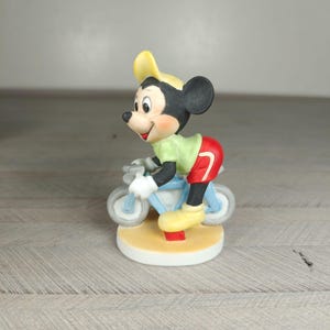 K&ouml;nnte beinhalten: Keramikfigur von Mickey Mouse auf einem hellblauen Fahrrad. Mickey tr&auml;gt eine gelbe M&uuml;tze, ein gr&uuml;nes Hemd, rote Shorts und gelbe Schuhe. Die Figur steht auf einem runden, beigen Sockel.