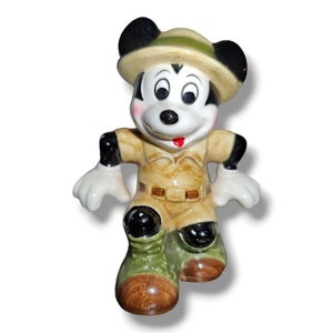 K&ouml;nnte beinhalten: Eine Keramikfigur von Micky Maus in Safari-Kleidung. Die Figur tr&auml;gt einen beigefarbenen Safari-Anzug, einen passenden Hut mit gr&uuml;nem Band und gr&uuml;ne Stiefel mit braunen Sohlen. Mickys schwarze Ohren und wei&szlig;en Handschuhe sind sichtbar.