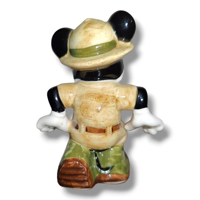 K&ouml;nnte beinhalten: Eine Keramikfigur von Micky Maus in Safari-Outfit. Die Figur ist von hinten zu sehen und tr&auml;gt ein beiges Hemd, gr&uuml;ne Hosen und einen Safarihut mit gr&uuml;nem Band. Die Figur hat schwarze Ohren, schwarze Handschuhe und wei&szlig;e Handschuhe.