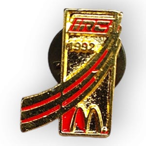 May include: A gold-coloured pin with red detailing and the text "IRC 1992". The pin features a stylized design with the McDonald's logo and a red ribbon-like element.