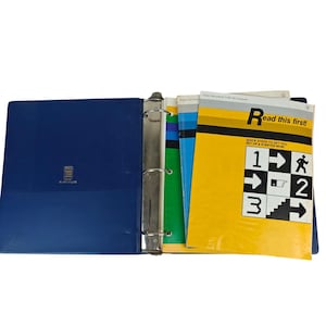 May include: A dark blue three-ring binder containing several manuals. The top manual features a yellow cover with the text Read this first! and a graphic grid showing numbered steps with arrows and icons for setting up a Texas Instruments TI-99/4A computer.