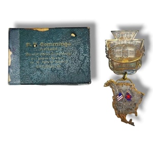 May include: A gold-toned metal badge with the text "B.P.R.T. Convention 1915" and a depiction of a building. The badge is attached to a silver-toned metal map of North America with two flags, one American and one Japanese.