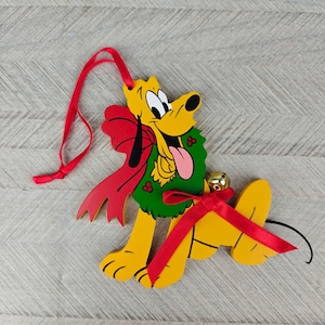 Op de afbeelding: Een gele Pluto honden kerstversiering met een rood lint en een groene krans. De versiering heeft een rood lint om op te hangen en een klein gouden belletje. De tong van de hond is roze en de strik is rood.
