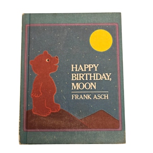 Puede incluir: Portada del libro infantil "Happy Birthday, Moon" de Frank Asch. Presenta un oso pardo, una luna amarilla y el título en texto blanco sobre un fondo azul estrellado. El libro tiene un borde turquesa.