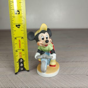 K&ouml;nnte beinhalten: Eine Keramikfigur von Mickey Mouse, die auf einem hellblauen Dreirad f&auml;hrt. Mickey tr&auml;gt ein gr&uuml;nes Hemd, eine blaue Hose, einen gelben Hut und einen roten Rucksack. Die Figur ist etwa 10 cm hoch.
